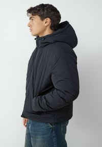 Schwarze Steppjacke mit Kapuze, aus leichtem Stoff, mit Reißverschluss und Seitentaschen. Glatte Textur mit wenigen Nähten.