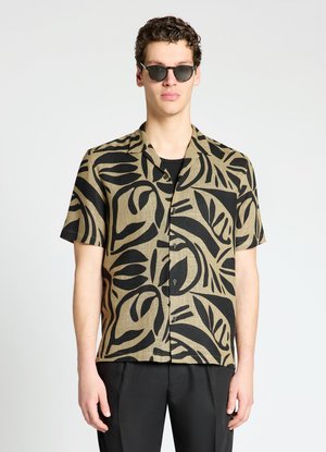 Uomo che indossa occhiali da sole neri e una camicia beige a maniche corte con audaci motivi astratti neri a forma di foglie, in piedi davanti a uno sfondo semplice.