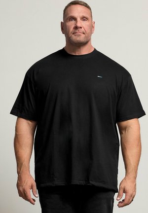 Muskuløs mand med kort hår og trimmet skæg står vendt fremad iført en løstsiddende sort t-shirt mod en ensfarvet baggrund.