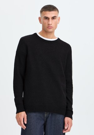 Jeune homme aux cheveux courts portant un pull noir par-dessus une chemise blanche et un jean bleu sur un fond clair uni.