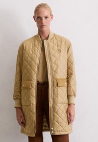 Femme portant un long manteau matelassé beige sur un pull et un pantalon marron, se tenant devant un mur blanc uni.