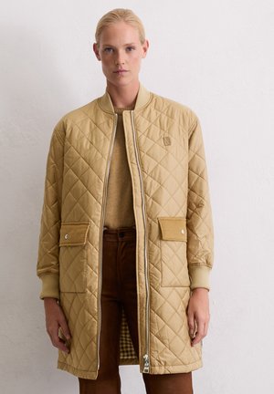 Vrouw in een tan gekwilt lang jack over een bruine trui en broek, staand tegen een plain witte muur.