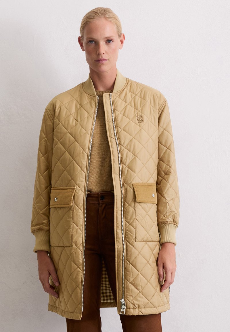 Femme portant un long manteau matelassé beige sur un pull et un pantalon marron, se tenant devant un mur blanc uni.