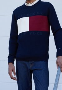 Pull côtelé bleu marine avec une bande horizontale rouge et blanche et le texte « HILFIGER NEW YORK ». Encolure arrondie, porté avec un jean en denim.