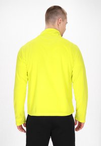 Pullover a maniche lunghe in tessuto giallo brillante con colletto alto e cuciture piatte; texture liscia, design aderente e pantaloni neri visibili in basso.