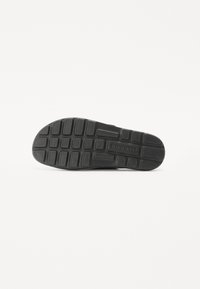 Semelle de chaussure en caoutchouc noir avec un motif texturé, présentant des crampons rectangulaires et carrés pour une meilleure adhérence. Le nom de la marque "bugatti" est visible.