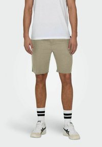 Pantaloni corti beige realizzati in tessuto testurizzato, caratterizzati da una vestibilità rilassata, che arrivano a metà coscia, abbinati a una maglietta bianca, calze a righe bianche e nere e sneakers.