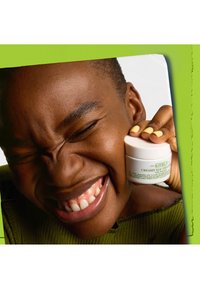 Kiehl's Creamy Eye Treatment-Glas, weißer Behälter mit grüner Schrift, glatte Oberfläche. Modell mit kurzem dunklen Haar, trägt ein strukturiertes grünes Oberteil.