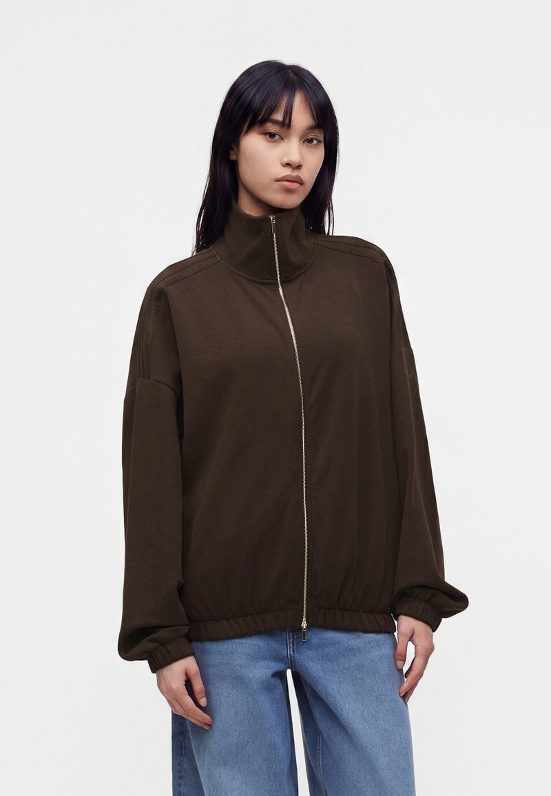 Femme aux cheveux noirs portant une veste marron foncé zippée avec un col haut et un jean bleu clair sur un fond uni clair.