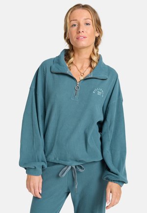 Teal velours pullover met een kwart-rits, geribbelde textuur, oversized mouwen en een rond logo op de borst. Gecombineerd met bijpassende broek.