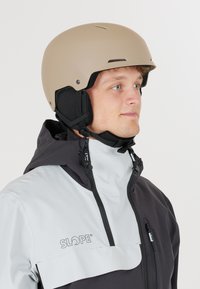 Jonge man met een beige helm en een zwart-witte Slope winterjas, kijkt iets naar rechts tegen een effen achtergrond.