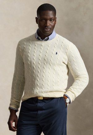 Homme portant un pull en maille torsadée crème sur une chemise rayée bleue, un pantalon bleu marine, une ceinture marron et une montre-bracelet, debout avec une main dans la poche.