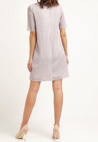 Robe grise claire à manches courtes, avec des plis verticaux, coupée au genou et à la coupe droite. Portée avec des chaussures à talons beiges.
