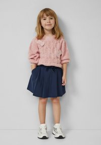 Rosa gestrickter Pullover mit strukturiertem Muster und Schleifendetails, kombiniert mit einem marineblauen Faltenrock. Weiße Sneakers komplettieren den Look.