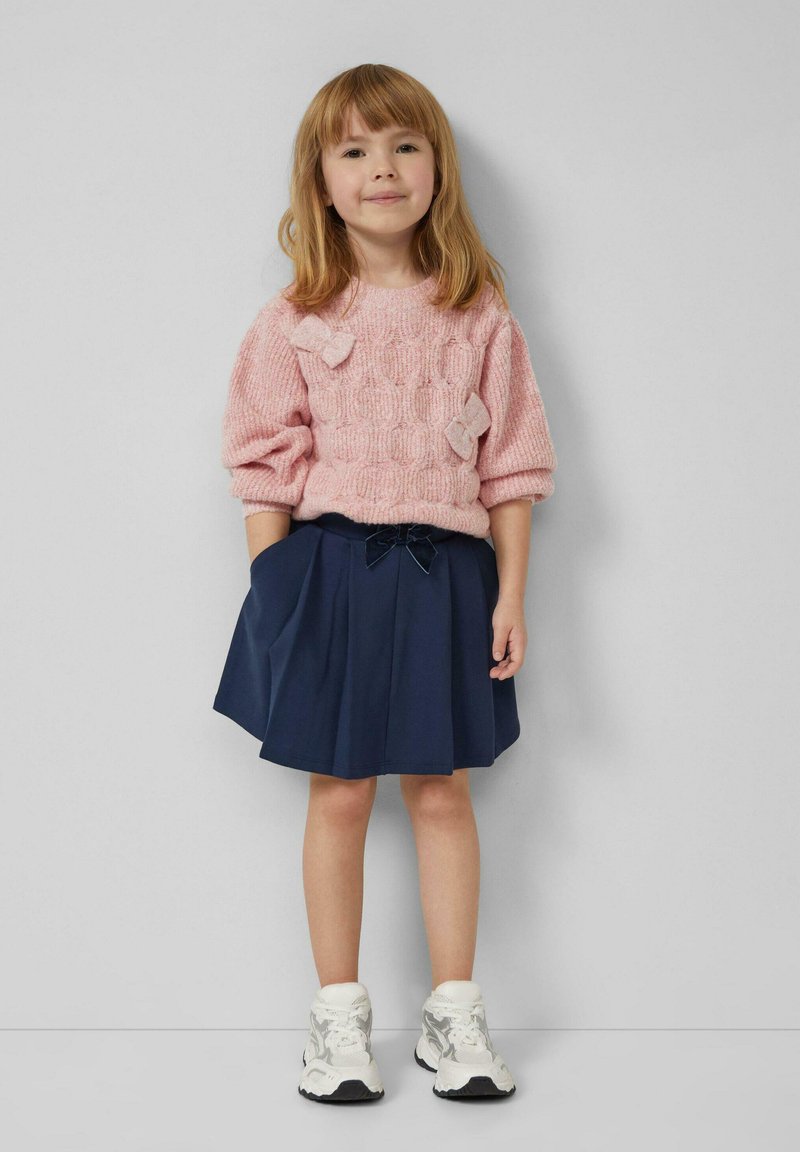 Rosa gestrickter Pullover mit strukturiertem Muster und Schleifendetails, kombiniert mit einem marineblauen Faltenrock. Weiße Sneakers komplettieren den Look.