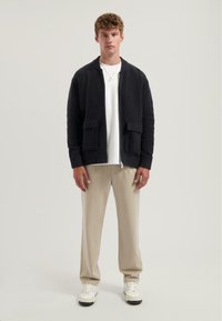 Černý pletený kardigan se dvěma předními kapsami, vroubkovanými manžetami a zipovým zapínáním, nošený přes bílé tričko a béžové kalhoty a bílé tenisky.