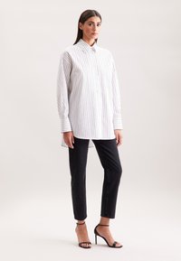 Vrouw in een wit, verticaal gestreept, lange mouwen shirt, zwarte slim fit jeans en zwarte hoge hakken, staande tegen een effen achtergrond.