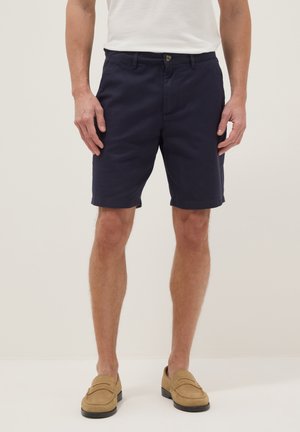Man met donkerblauwe shorts, beige loafers en een wit overhemd, staand tegen een effen lichte achtergrond.