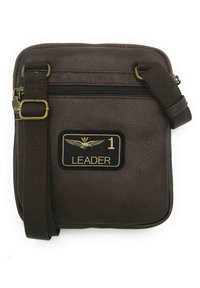Borsa a tracolla in eco-pelle marrone con chiusura a zip, tracolla regolabile e una patch nera con testo dorato e un emblema.