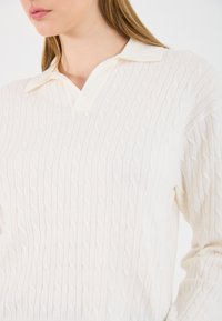 Κρεμ μάλλινη πουλόβερ με υφή καμπάνας, που διαθέτει μ�αλακό γιακά και διακριτικό σχέδιο V στον λαιμό.