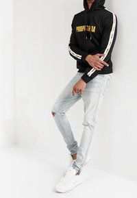 Schwarzer Kapuzenpullover mit "PROPAGANDA" in Gelb, weiß gestreifte Ärmel; hell bleached Jeans mit einem gerissenen Knie; weiße Sneakers.