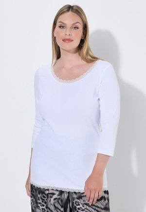 Donna con indosso una maglia bianca a manica lunga con bordo in pizzo e pantaloni con stampa animalier bianco e nero, in piedi davanti a uno sfondo chiaro e semplice.