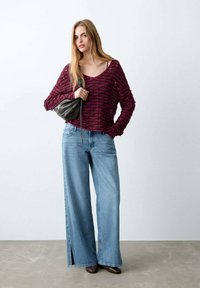 Pulover tricotat bordo cu decolteu larg, asortat cu blugi evazați bleu deschis, cu tăieturi laterale. Ține o geantă neagră, poartă pantofi maro.