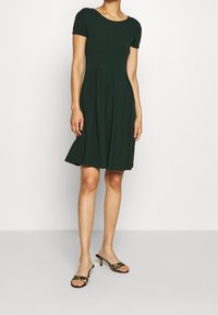 Robe noire verdâtre jusqu'aux genoux avec des manches courtes, corsage ajusté et jupe évasée. Portée avec des sandales à talons imprimé léopard.