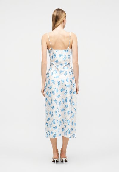 Γυναίκα που στέκεται με την πλάτη γυρισμένη, φορώντας λευκό midi φόρεμα τύπου slip με μπλε λουλουδάτο σχέδιο και λεπτές τιράντες στους ώμους, συνδυασμένο με λευκά τακούνια.