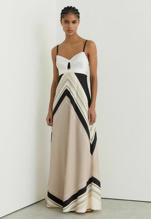 Kvinde med fletninger iført en lang, ærmeløs kjole med hvid top, sorte stropper og nederdel med geometrisk mønster i sort, beige og hvid.