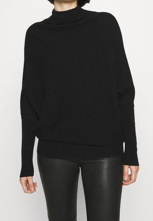 Personne portant un pull à col roulé noir ample et un pantalon en cuir noir, debout devant un fond clair uni.