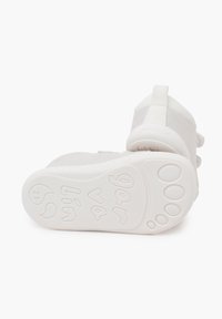Zapatos infantiles blancos con un upper de tela suave, diseño curvado y suela de goma texturizada que presenta un patrón de huellas de patas y una marca en relieve.