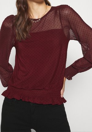 Vrouw draagt een doorschijnende bordeauxrode blouse met lange mouwen, met een gestippelde textuur, geplooide zoom en manchetdetails, gecombineerd met een zwarte broek.