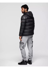 Giacca a vento nera con cappuccio, caratterizzata da un design trapuntato. Abbinata a pantaloni jogger grigi con tasche con zip e sneakers nere.