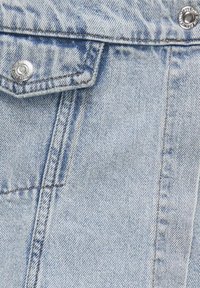 Giacca in denim azzurro chiaro con taschino sul petto, bottoni a pressione argentati e dettagli di cucitura visibili. Il tessuto ha una consistenza morbida con una finitura sbiadita.