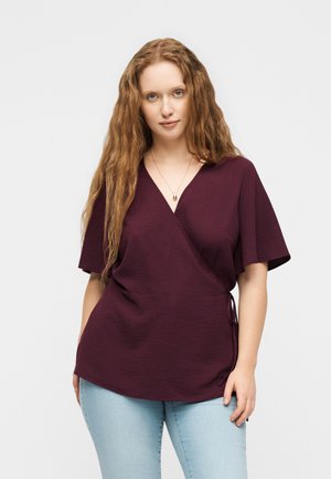 Γυναίκα με μακριά κυματιστά κόκκινα μαλλιά, ντυμένη με ένα μωβ σκουρόχρωμο τοπ τύπου wrap με κοντά μανίκια και ανοιχτό μπλε τζιν, στέκεται μπροστά σε απλό φόντο.