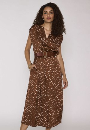 Femme aux cheveux bouclés portant une robe midi marron à pois cintrée par une large ceinture en cuir, debout avec une main dans la poche.