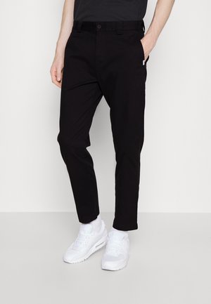 Schwarze, maßgeschneiderte Chinos mit schmaler Passform, aus festem Stoff, mit einem Knopfverschluss, seitlichen und Rückentaschen. Kombiniert mit weißen Sneakers und Socken.