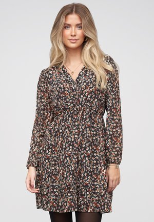 Jonge vrouw met lang blond haar, gekleed in een zwarte jurk met bloemenprint en lange mouwen, met een gouden ketting, staand tegen een effen lichte achtergrond.