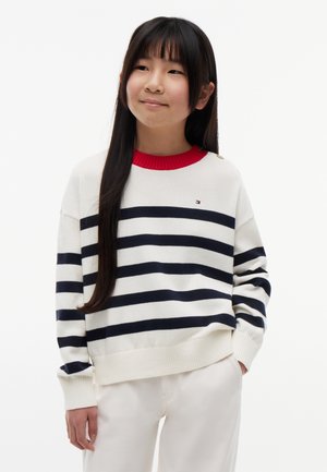 Ragazza con lunghi capelli scuri che indossa un maglione bianco con righe orizzontali blu navy e colletto rosso, in piedi con le mani nelle tasche dei pantaloni bianchi.
