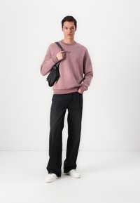 Helles rosa Sweatshirt mit gerippten Bündchen, kombiniert mit schwarzen weit geschnittenen Jeans und weißen Sneakern. Eine kleine schwarze Umhängetasche wird getragen.