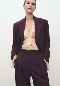 Blazer y pantalones ajustados burdeos combinados con una blusa crema con volantes, adornada con un delicado collar de perlas y texturas suaves y fluidas.