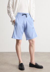 Lyseblå shorts laget av myk stoff, med elastisk midjebånd og svart snor, samt sidelommer. Brodert logo detaljer.