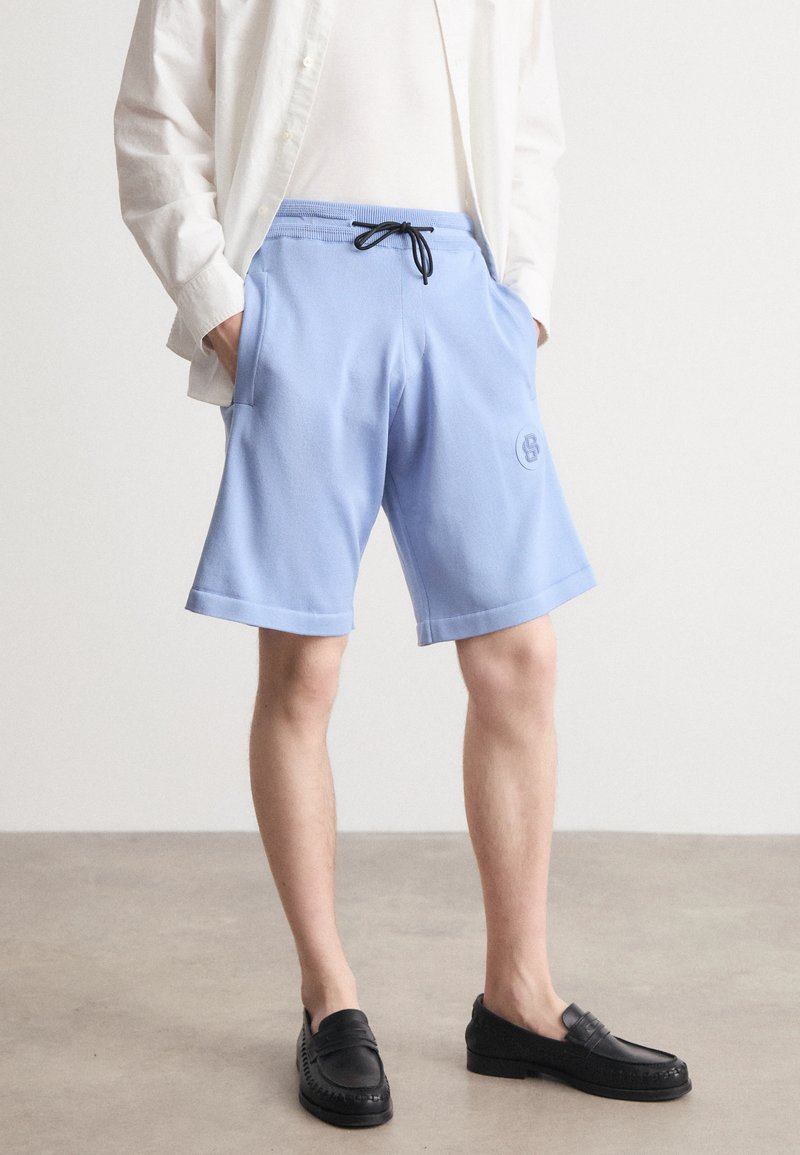 Lyseblå shorts laget av myk stoff, med elastisk midjebånd og svart snor, samt sidelommer. Brodert logo detaljer.