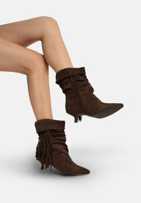 Brązowe botki z zamszu o luźnym fasonie, z ostrym noskiem i ozdobnymi frędzlami. Posiadają zamek błyskawiczny po boku oraz niewielki obcas.