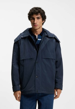 Ung mand iført en mørkeblå hættetrøje over en mørkeblå stribet sweater og blå jeans, stående mod en ensartet hvid baggrund.