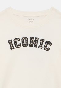 Κρεμ βαμβακερό μπλουζάκι με τη λέξη «ICONIC» σε έντονα μαύρα γράμματα με υφή λεοπάρ. Στρογγυλή λαιμόκοψη, κοντά μανίκια.