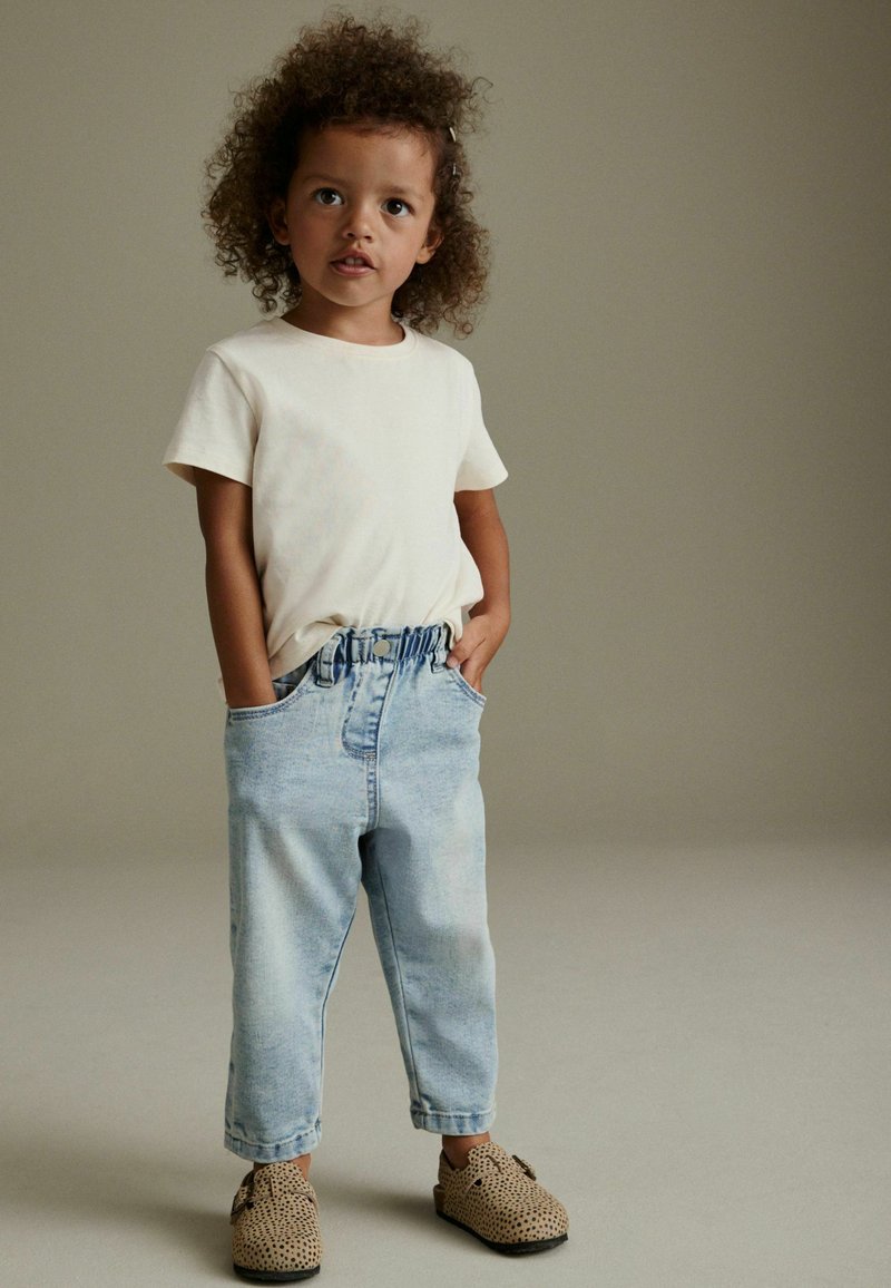 Kleines Kind mit lockigem Haar steht mit den Händen in den Taschen, trägt ein weißes kurzärmeliges Hemd, hellblaue Jeans und braune gepunktete Slipper.