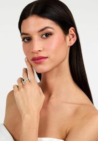 Anello d'argento con una pietra verde ovale, incastonata al centro, circondata da piccole pietre trasparenti. Indossato su una mano appoggiata vicino al viso.
