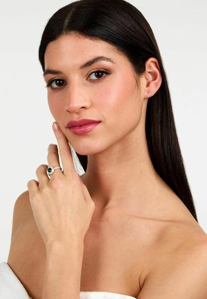 Anello d'argento con una pietra verde ovale, incastonata al centro, circondata da piccole pietre trasparenti. Indossato su una mano appoggiata vicino al viso.